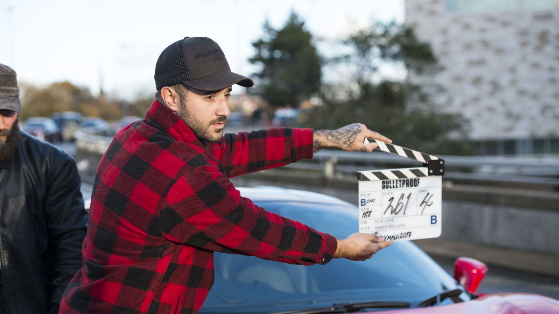 A man wearing a red checked shirt and baseball cap holds a clapper board in front of a sports car