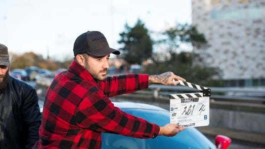 A man wearing a red checked shirt and baseball cap holds a clapper board in front of a sports car