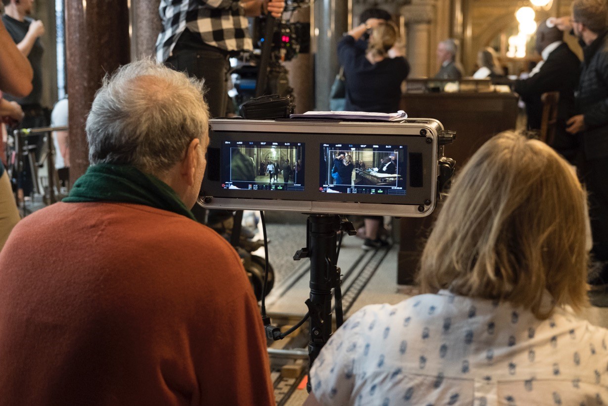 Two directors watch a scene playing out on a small screen while actors and crew set up a courtroom scene in the background