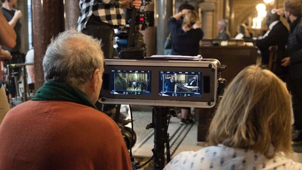 Two directors watch a scene playing out on a small screen while actors and crew set up a courtroom scene in the background