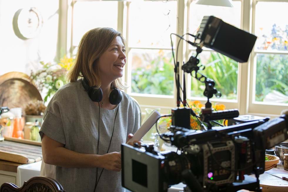 A woman wearing headphones stands behind a camera giving instructions