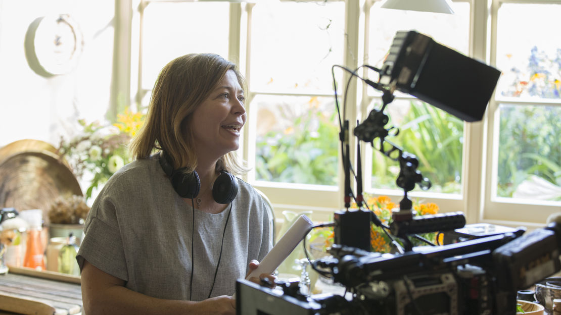 A woman wearing headphones stands behind a camera giving instructions