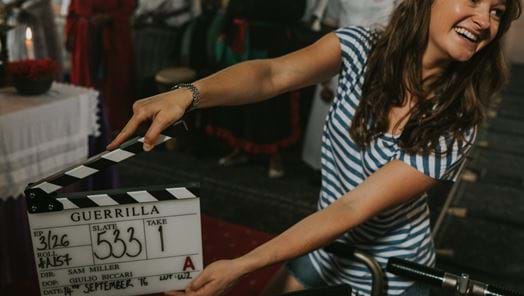 Behind-the-scenes image of a woman smiling while holding a clapper board on the set of the movie Guerilla