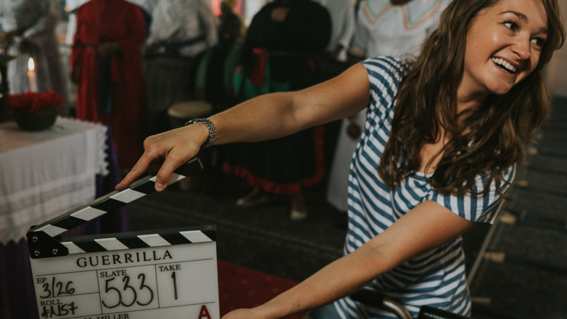 Behind-the-scenes image of a woman smiling while holding a clapper board on the set of the movie Guerilla