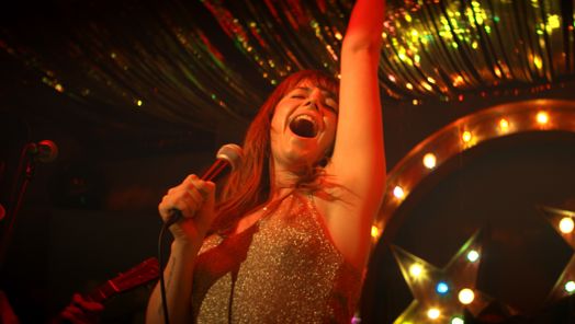 Wild Rose production shot of actress Jessica Buckley as the character Rose-Lynn signing into a microphone on a stage with star lights behind