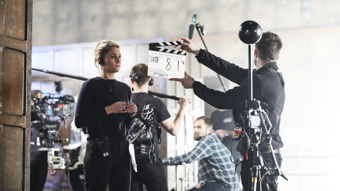 Man with clapper board stands in front of a woman wearing a headset with a 360 camera rig in the foreground