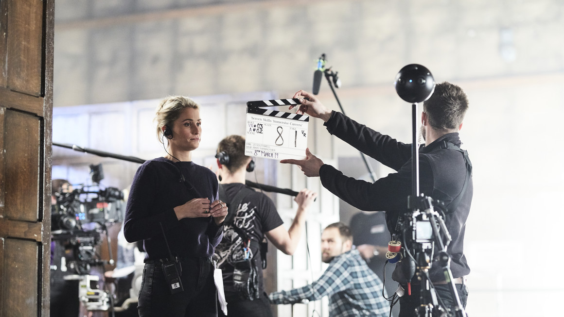 Man with clapper board stands in front of a woman wearing a headset with a 360 camera rig in the foreground