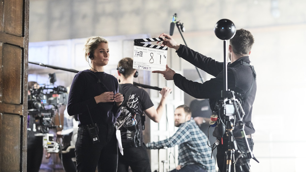 Man with clapper board stands in front of a woman wearing a headset with a 360 camera rig in the foreground