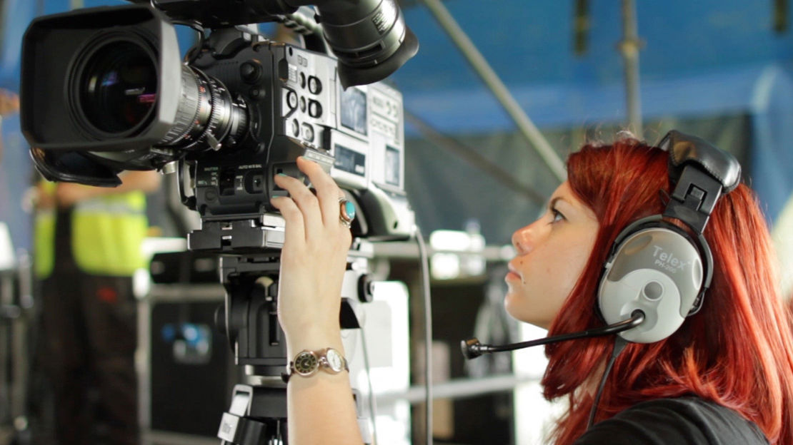 A young person wearing headphones inspects a TV camera on a tripod