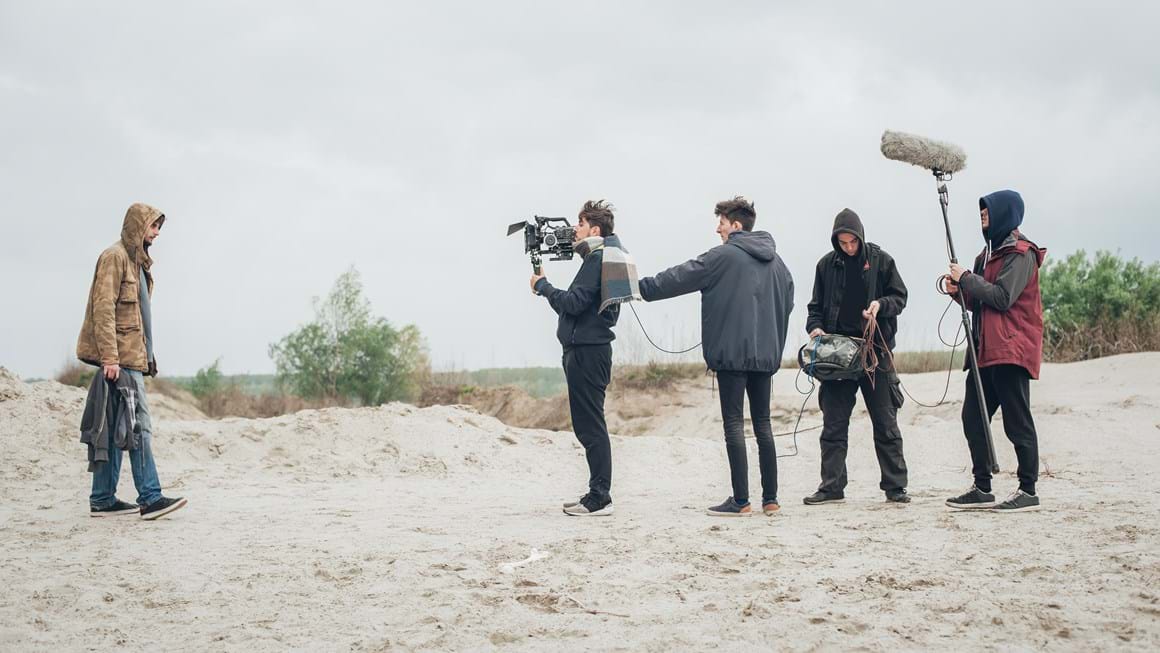 Camera and sound crew film a man in a hooded coat and trainers walking on a beach
