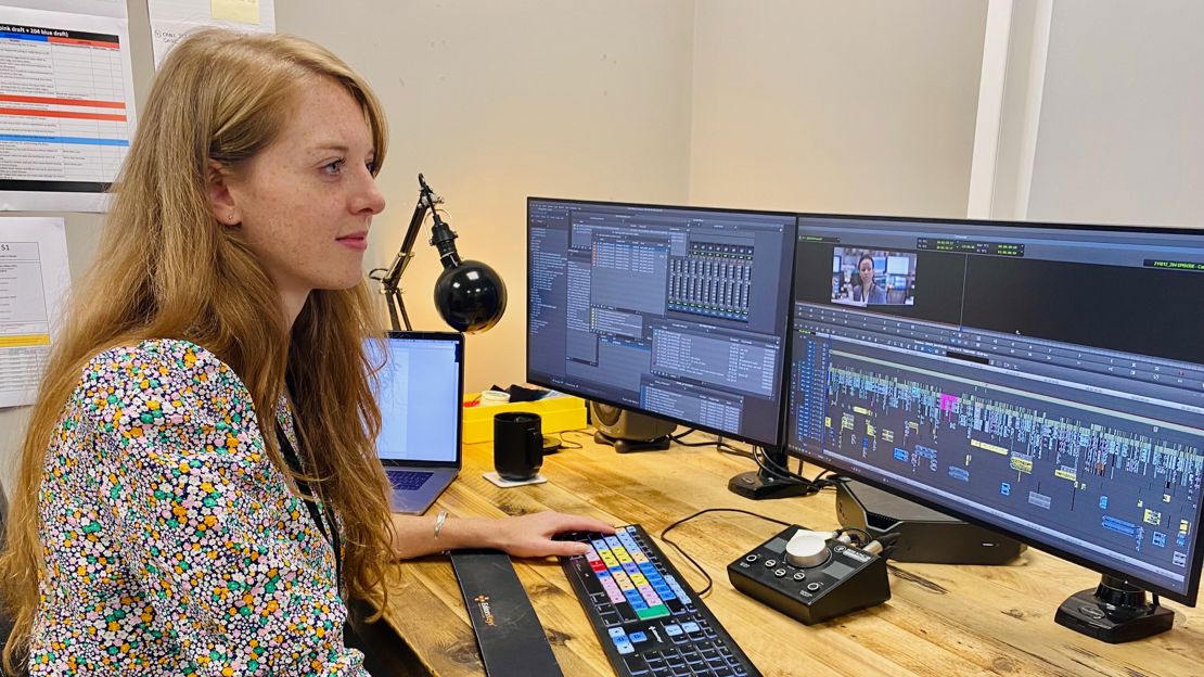 A woman sits at a desk in front of a set of computer monitors editing a sequence in a video editing software.