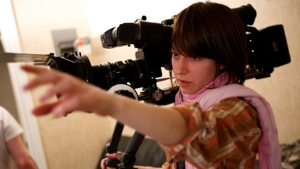 Close up of young person holding a large camera on their shoulder while reaching out with the other arm