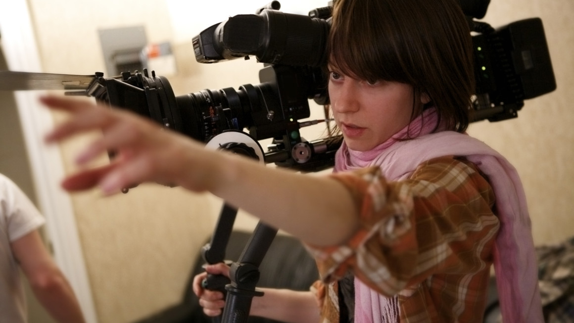 Close up of young person holding a large camera on their shoulder while reaching out with the other arm