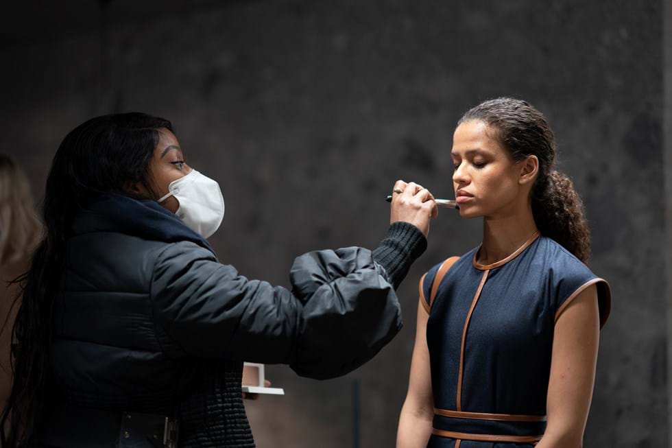 A make-up artist wearing a facemask applies foundation to an actor, who is standing with her eyes closed