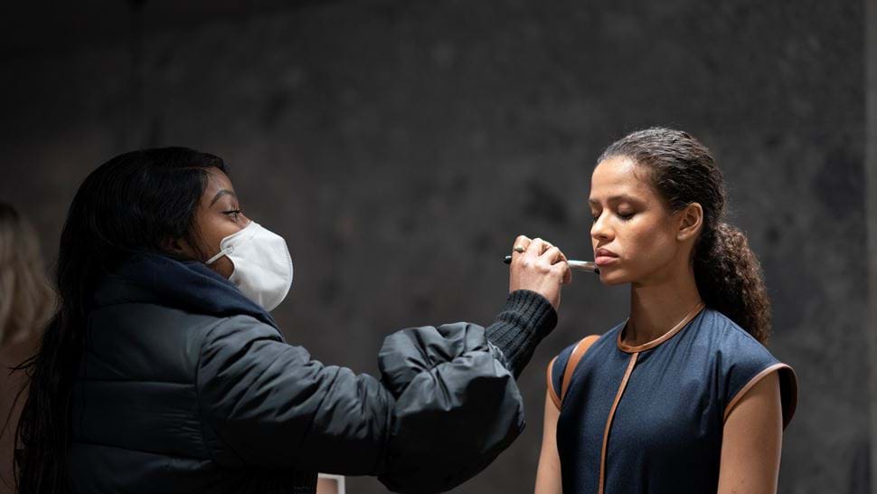 A make-up artist wearing a facemask applies foundation to an actor, who is standing with her eyes closed