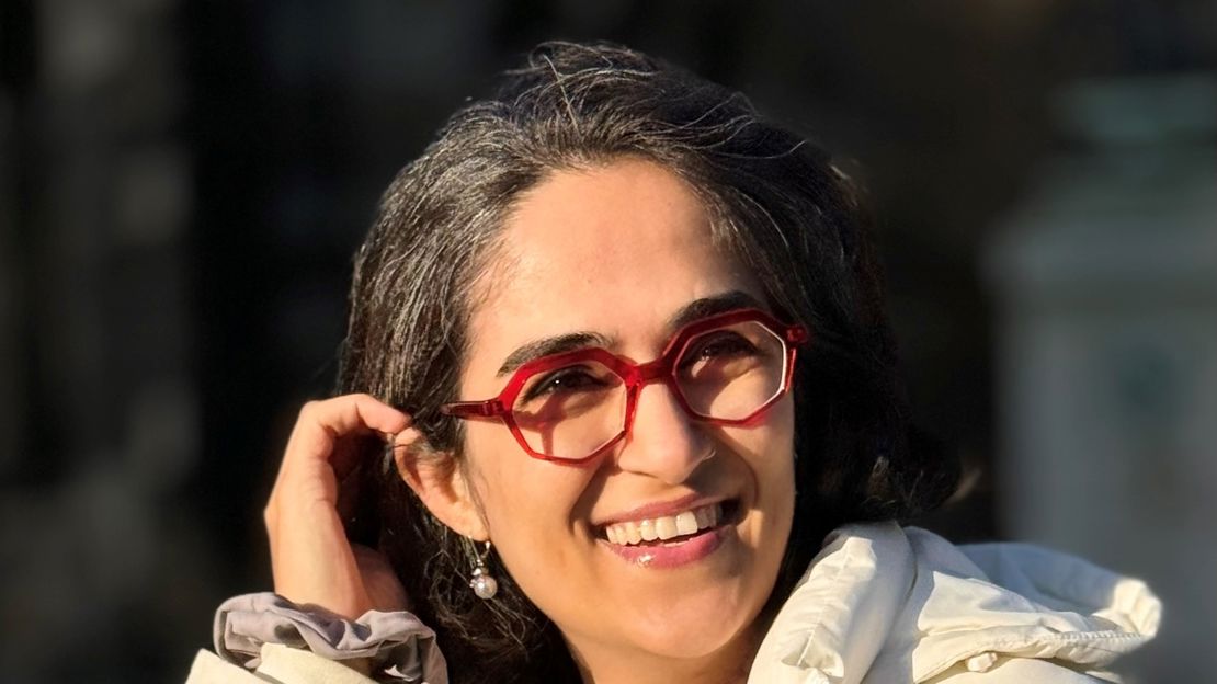 Elahe Esmaili stands smiling in a white coat with her arm raised to push back hair