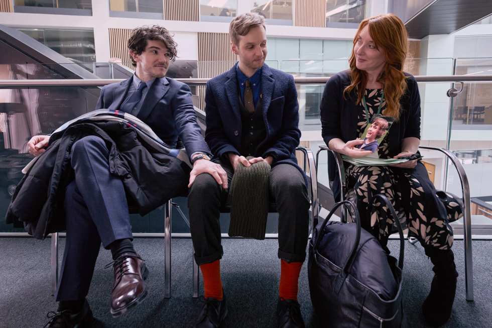 A man puts his hand on another man's knee as the pair sit next to a smiling woman