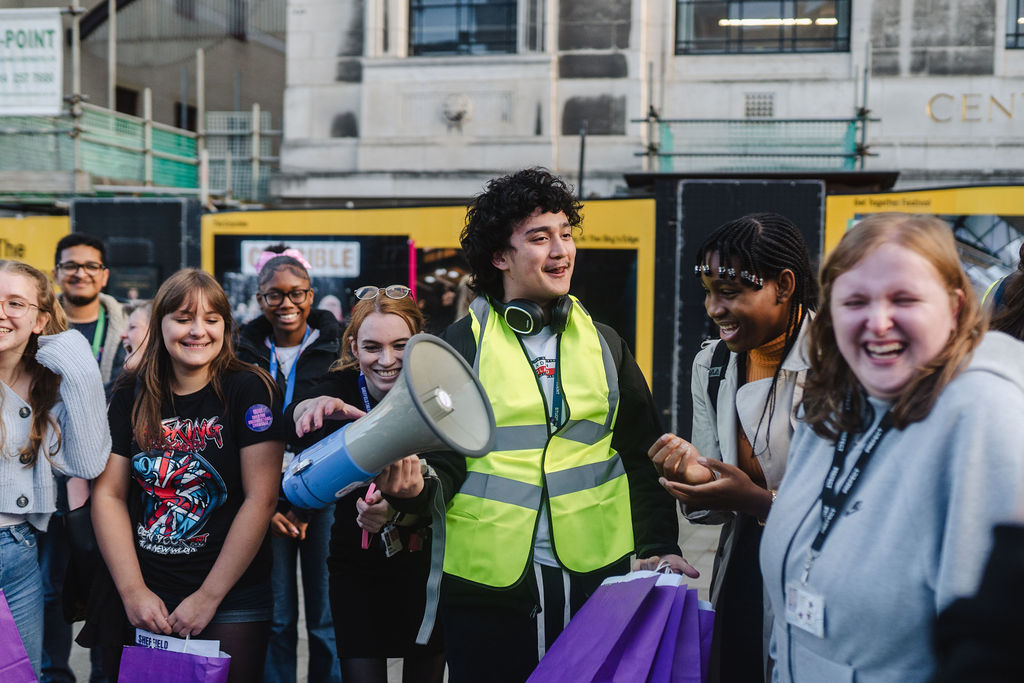A group of young people stand together outside laughing and smiling. One person wearing a Hi-vis jacket is centred in the middle of the group holding a megaphone