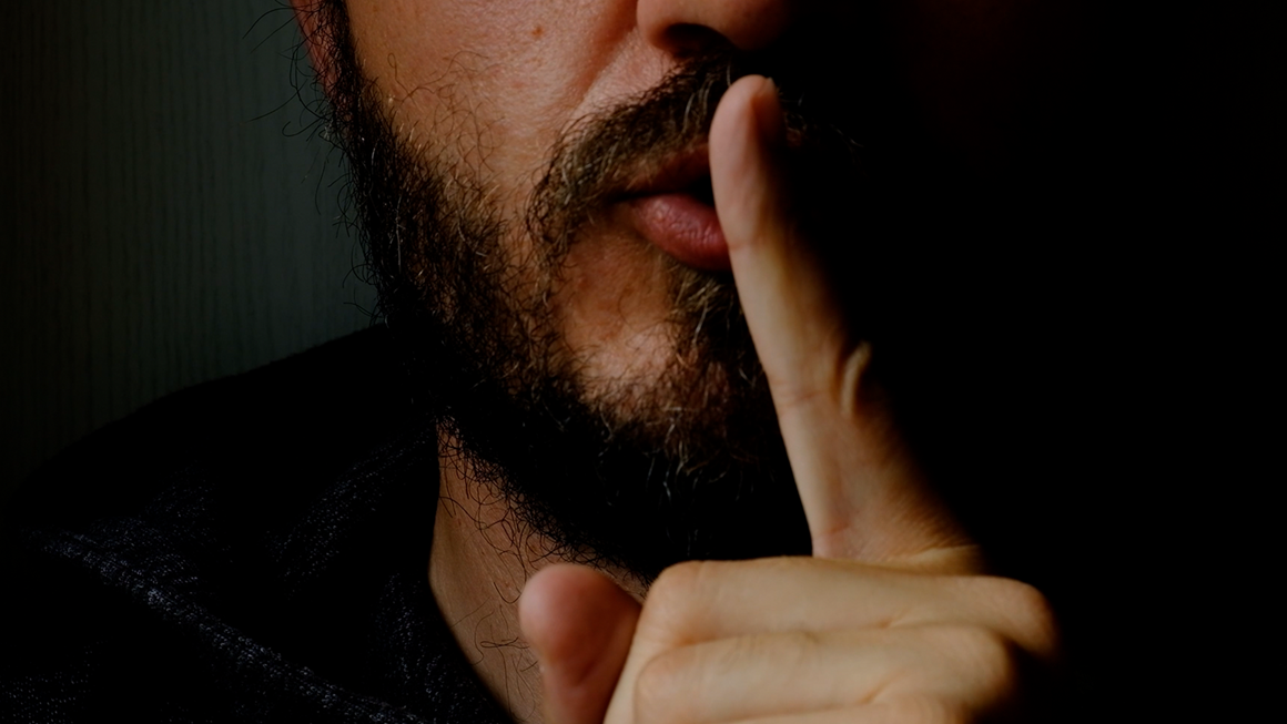 Close up of a bearded man holding his index finger in front of his mouth in a silence motion