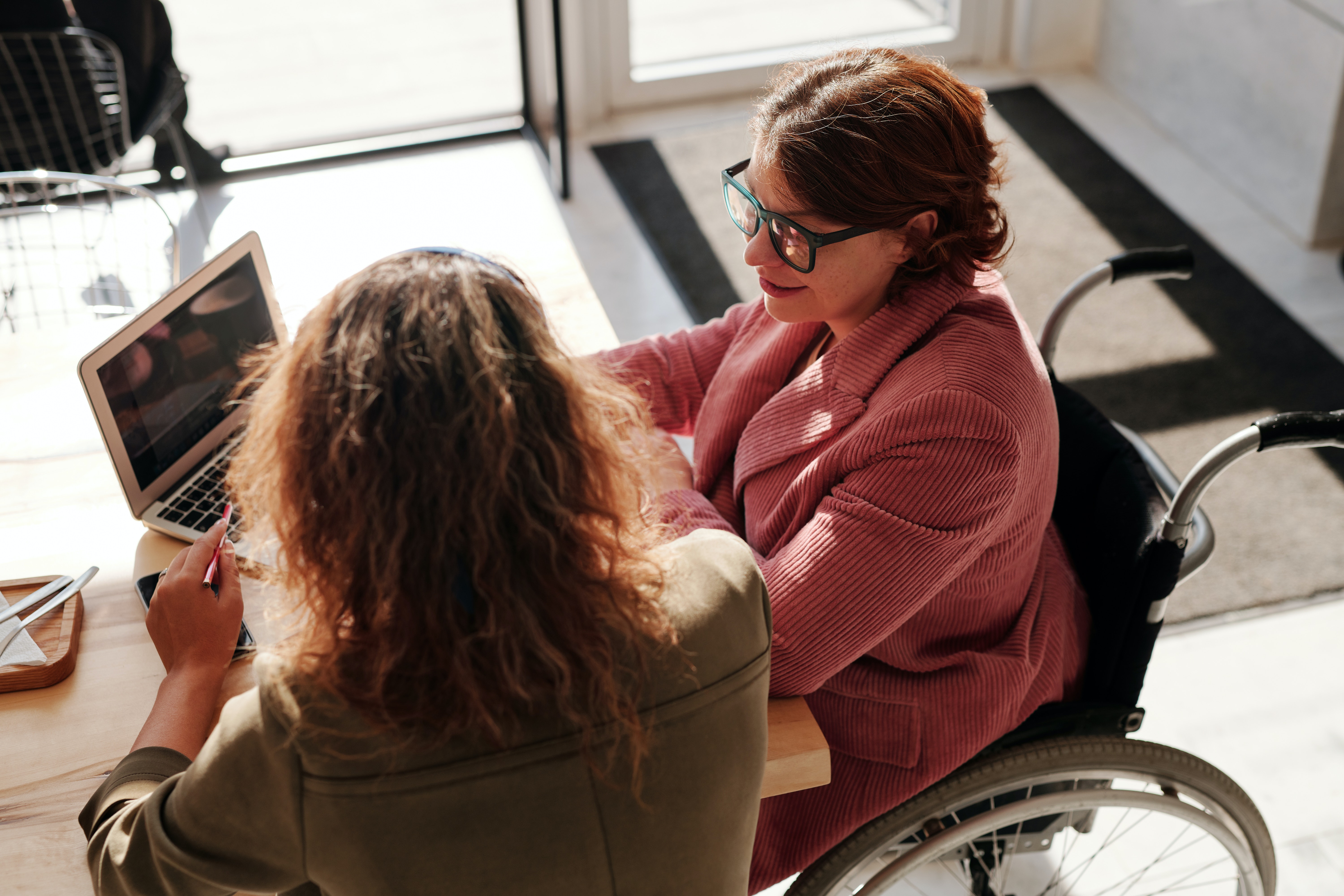 Two women, one in a wheelchair, sit at a desk looking at a laptop