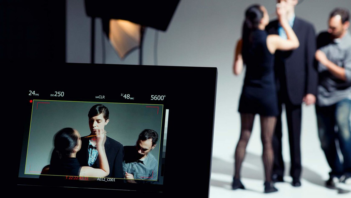 Camera viewer in the foreground showing a person applying make up to an actor and a man adjusting his costume, with the three people visible in the background