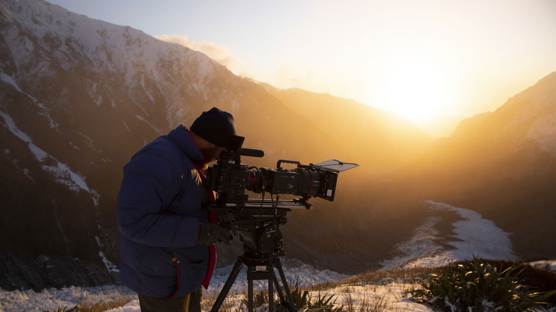 Camera operator is shooting a scene in the snow, wearing a heavy jacket and a hat.