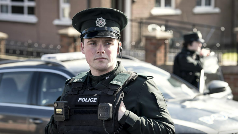 Production still of a police officer in front of a police car in Blue Lights