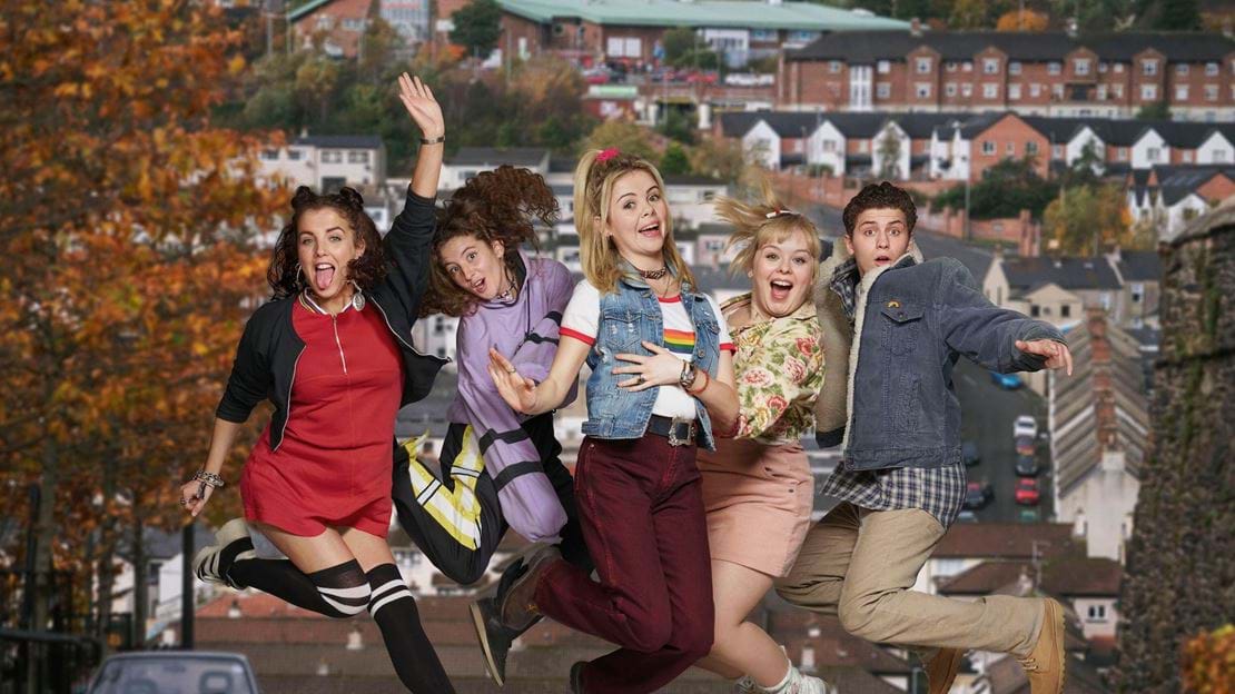 Cast of the Derry Girls jumping while smiling and waving in the middle of a street