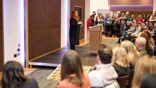 Michaela Mccaffrey stands at a podium in front of a seated audience giving a presentation