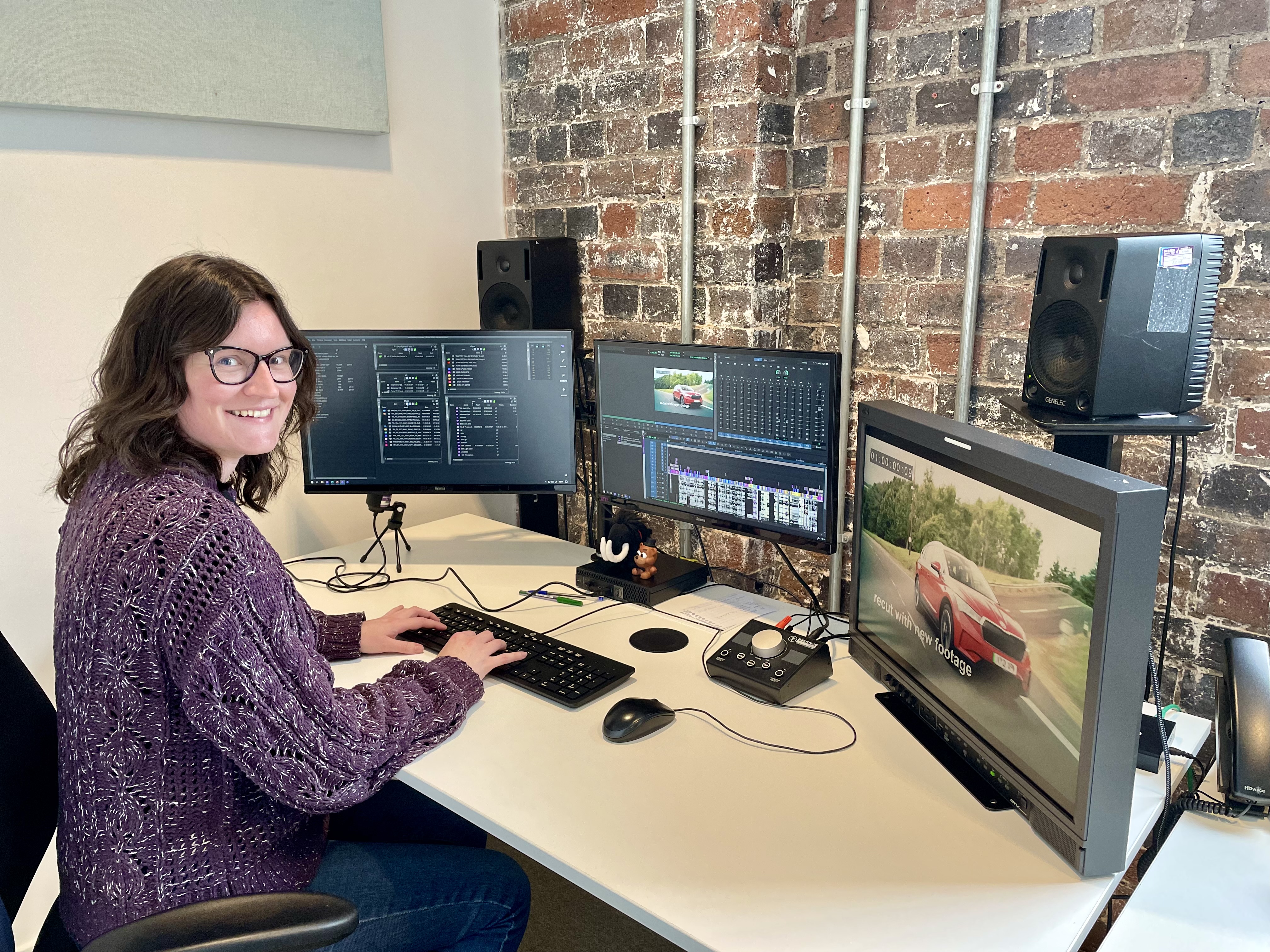 Kate Burley sits at an edit desk in front of three large screens and a set of speakers