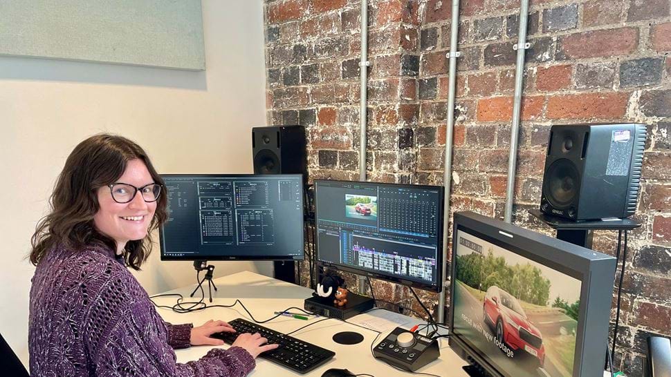 Kate Burley sits at an edit desk in front of three large screens and a set of speakers