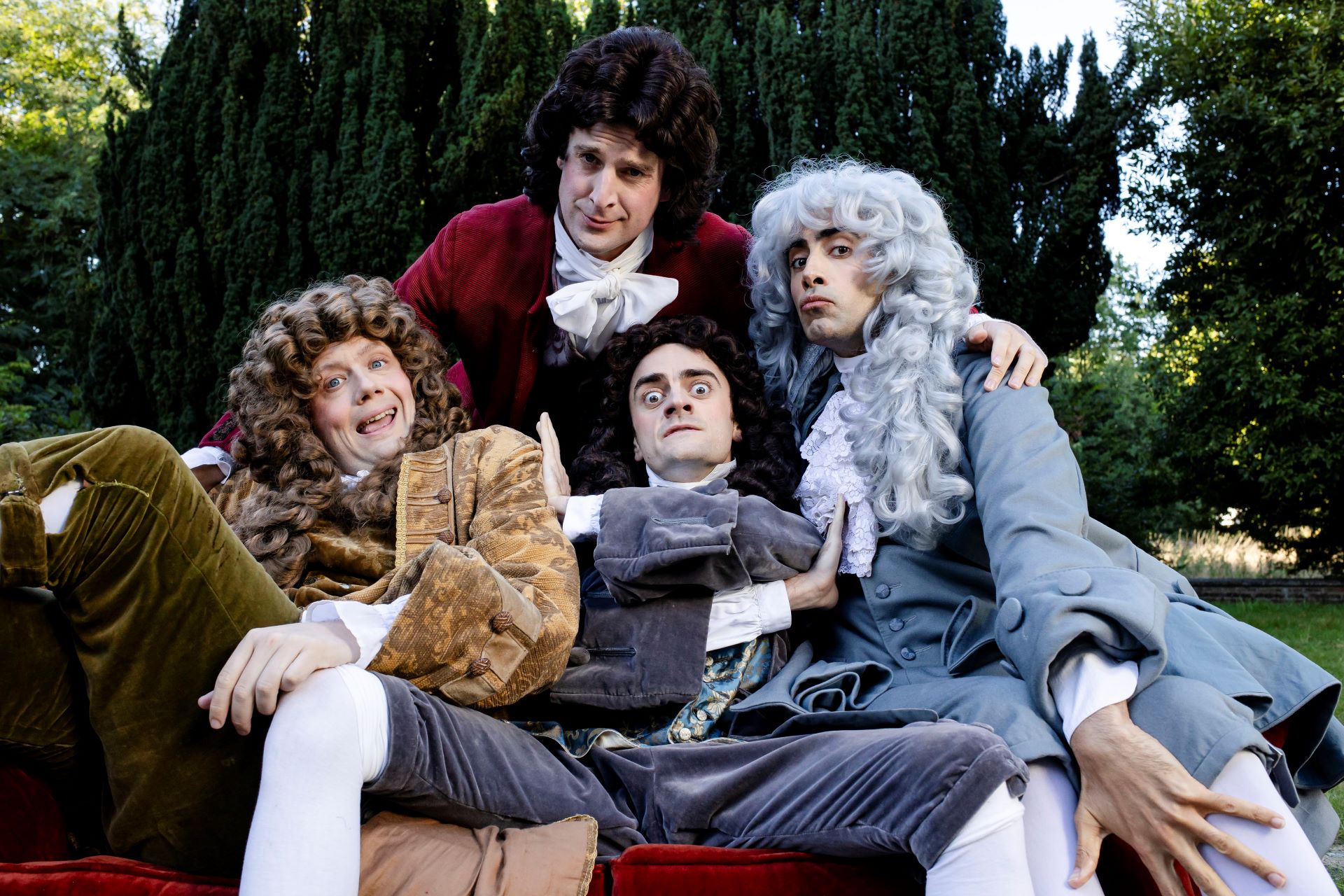 Four men sit posing together on a sofa dressed in period attire and elaborate wigs