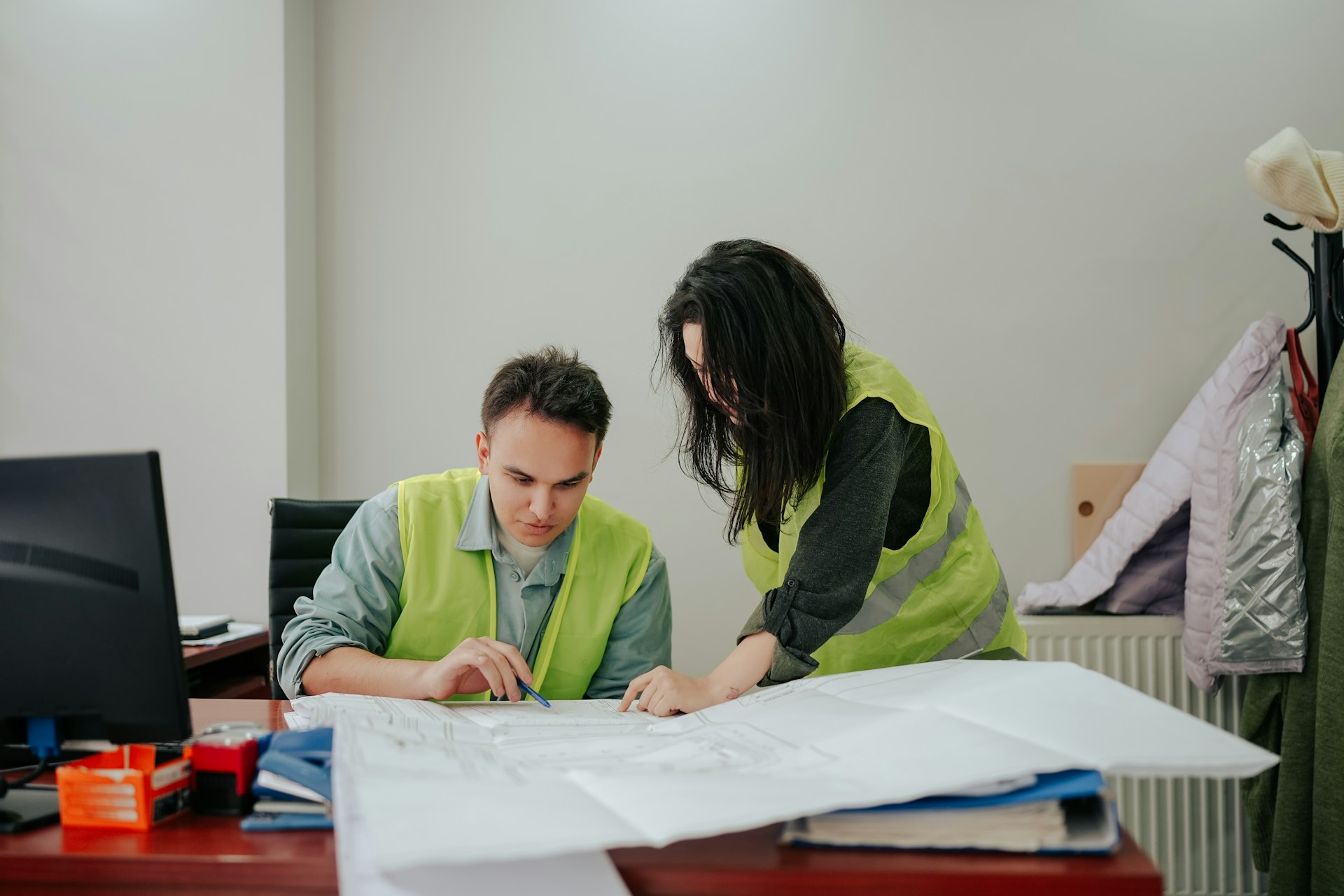 Two people wearing hi-vis jackets look over a large sheet of paper at a desk