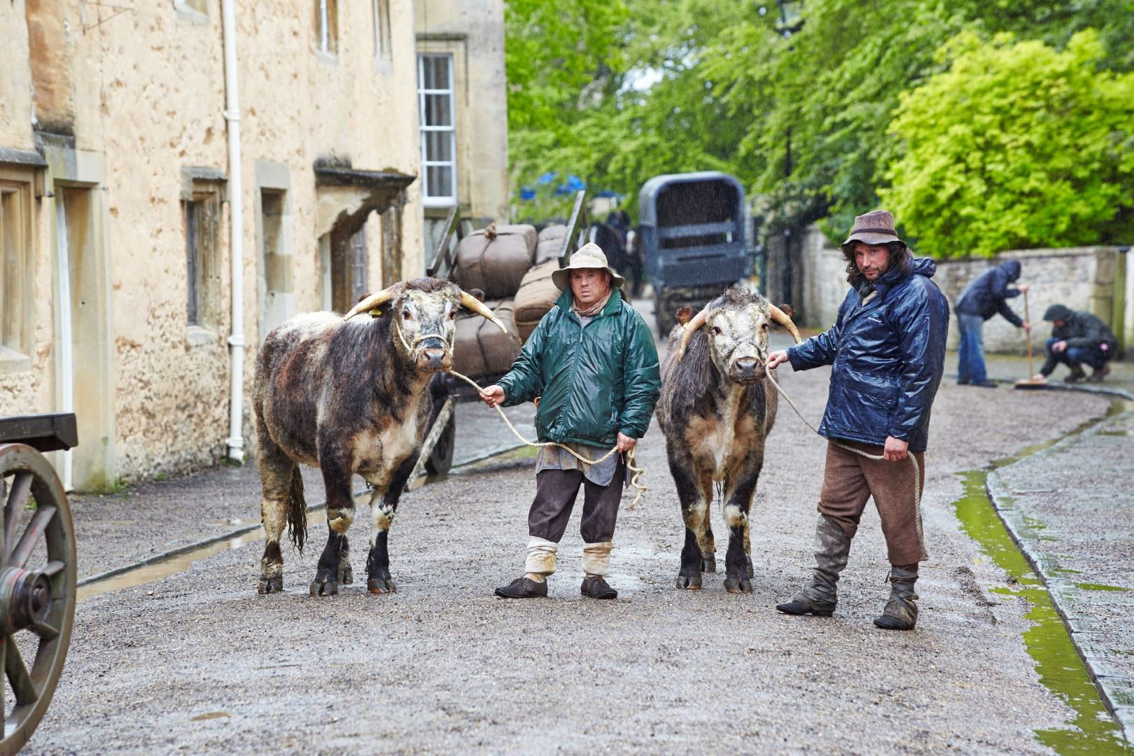 Two men dressed in waterproof clothing stand on the set of a period drama. Both are holding onto a rope attached to a cow.