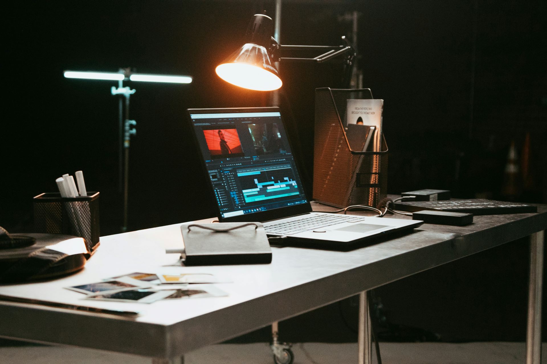 A laptop is open on a desk surrounded by books, papers and equipment. An overhead lamp shines on the screen which is open to a video editing software.