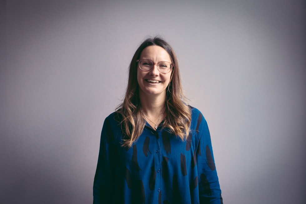 Lucy Watt, wearing a blue shirt and glasses, smiles for a posed photo in front of a grey background