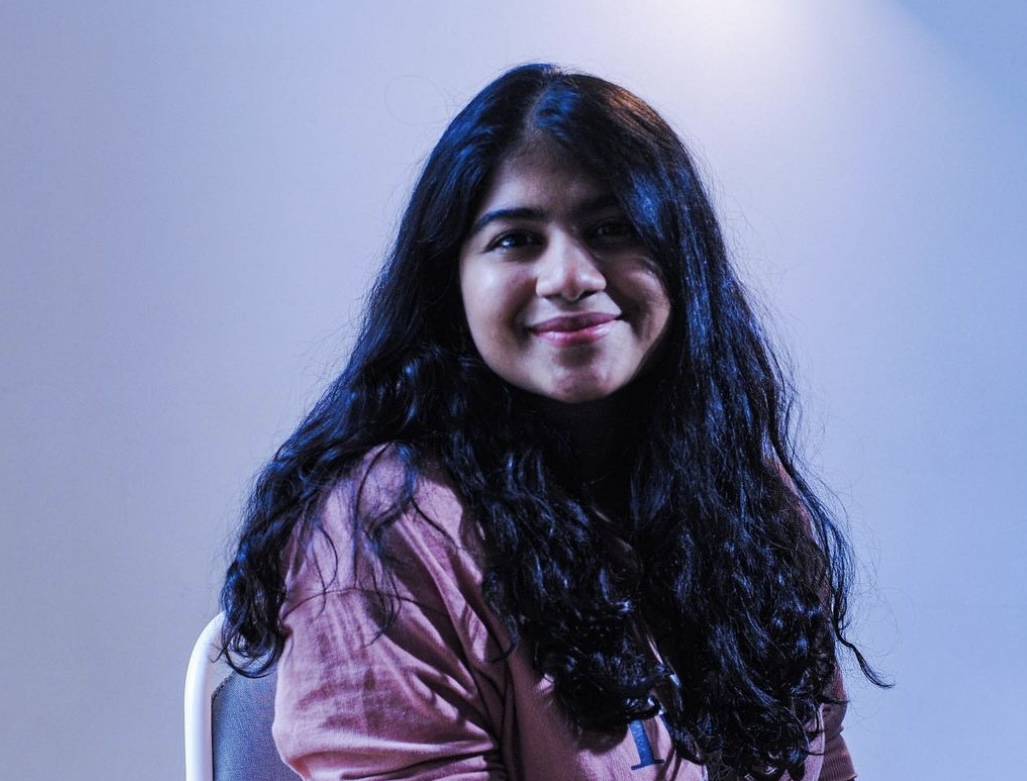 A headshot of Sanjoli Malani smiling towards the camera in front of a pale plain background