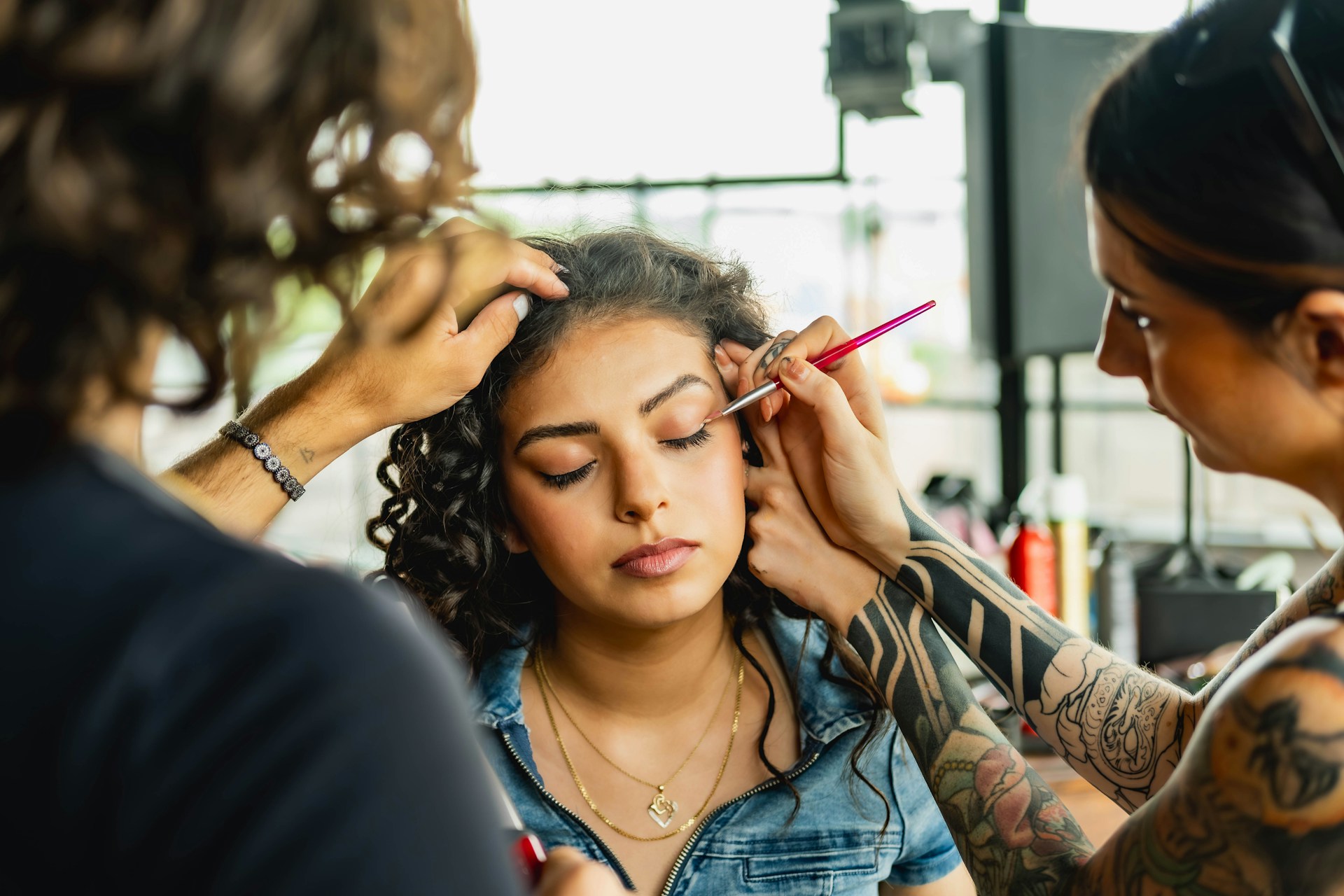 A woman sits in a chair while hair and makeup artists work with her