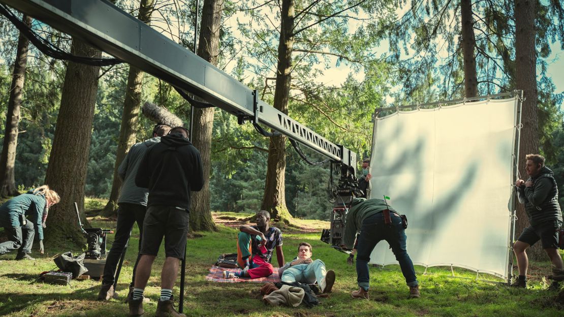 On location for Sex Education season 3, two actors lounge on picnic blankets in a forest in front of a large white screen and a camera boom