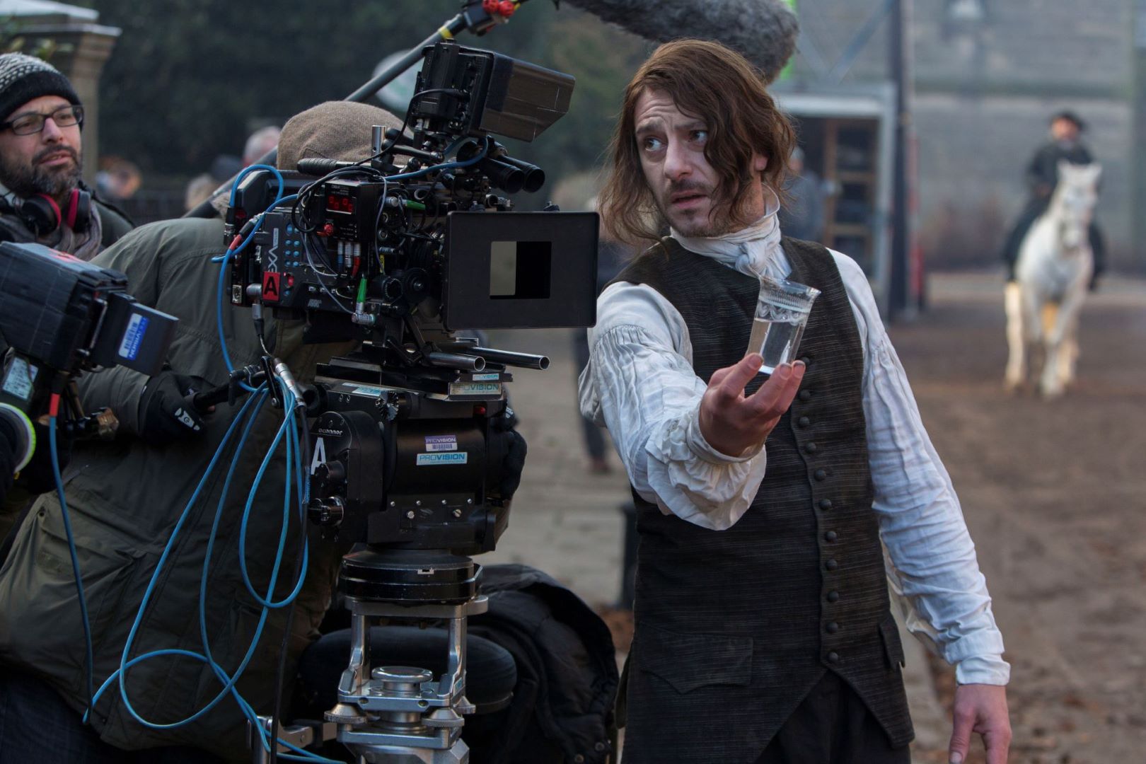 On the set of period drama, a man stands holding a glass. He looks towards a large film camera.