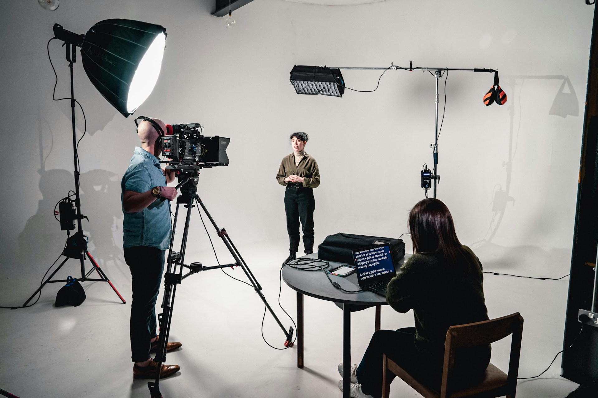 An indoor film shoot with a cameraman operating equipment and a person sat with a teleprompter on a table