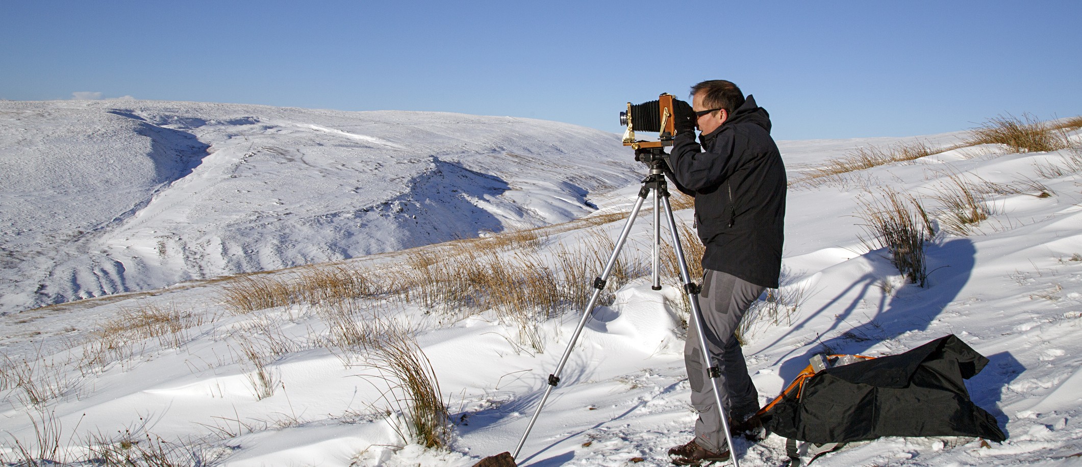 A man in protective weather gear stands in a snowy landscape looking through a large old-fashioned camera on a tripod
