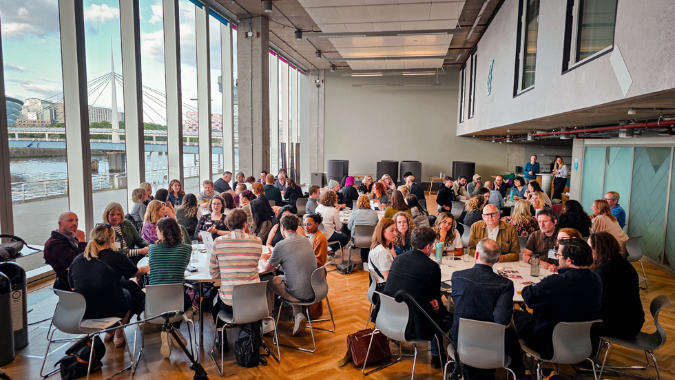 A range of people sit around several large round tables in a conference room overlooking a river