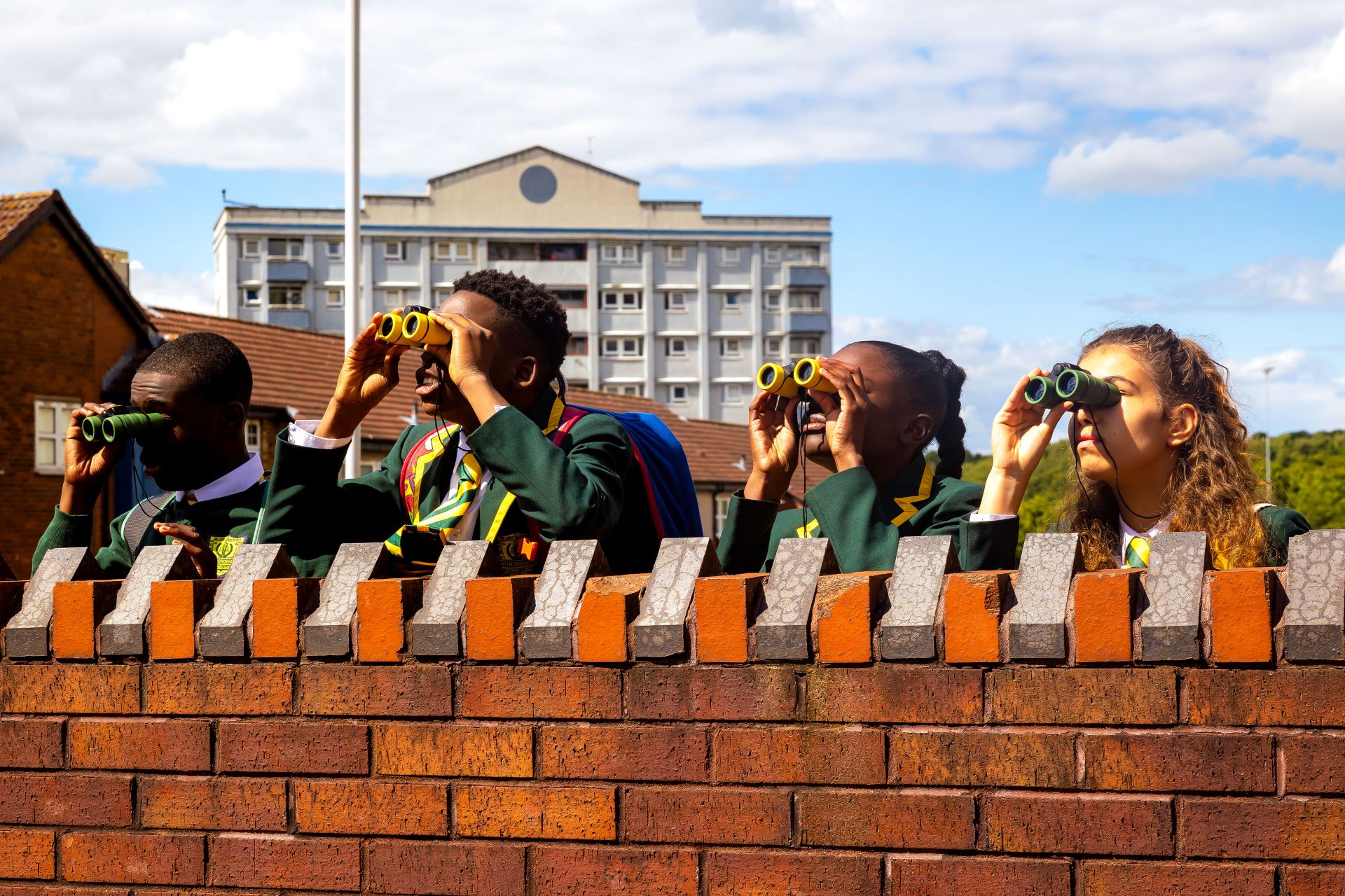 Four children in school uniform stand looking over a wall with a pair of binoculars each