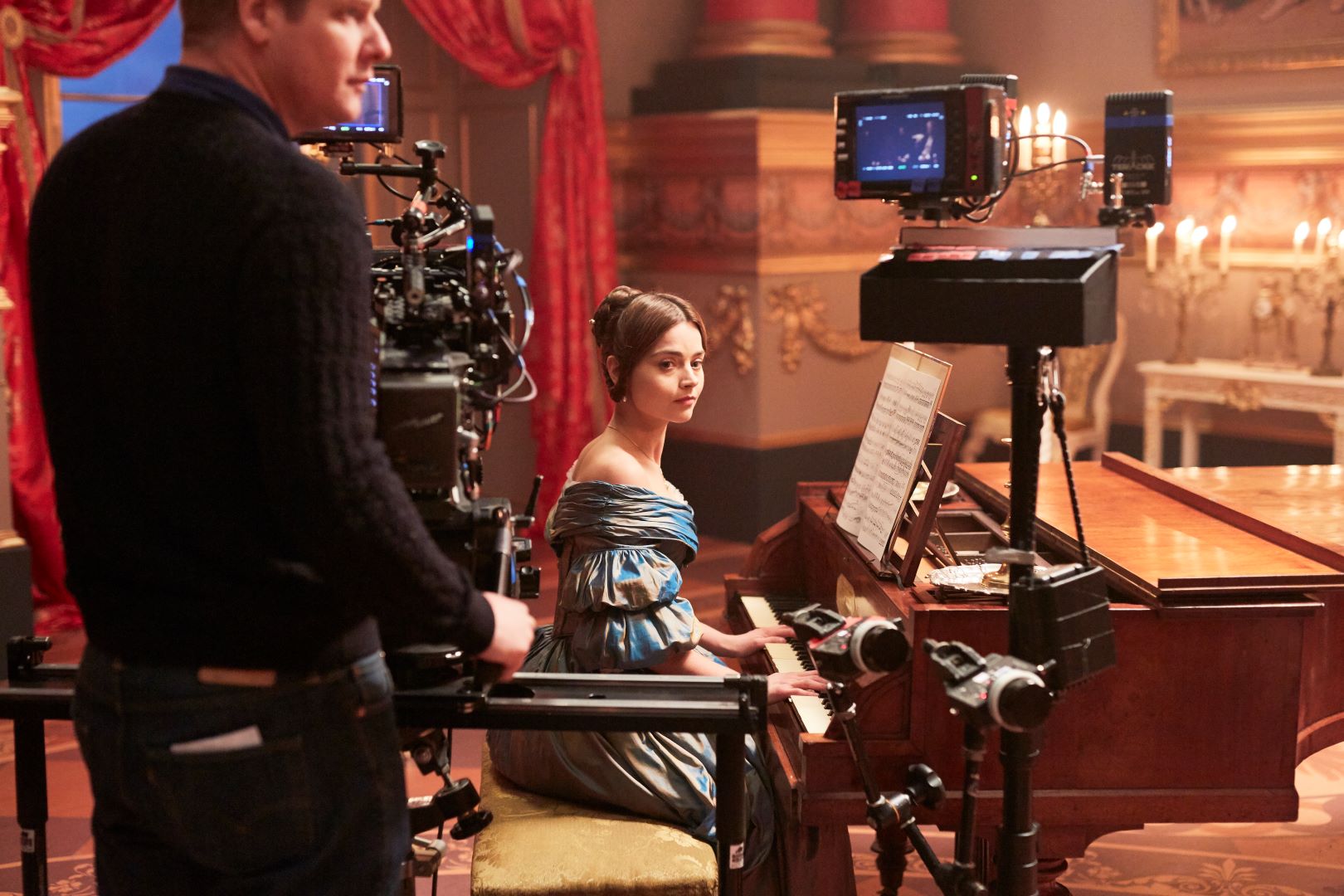 An actress sits at a piano on the set of a period drama. In the foreground, a camera operator stands monitoring camera equipment.