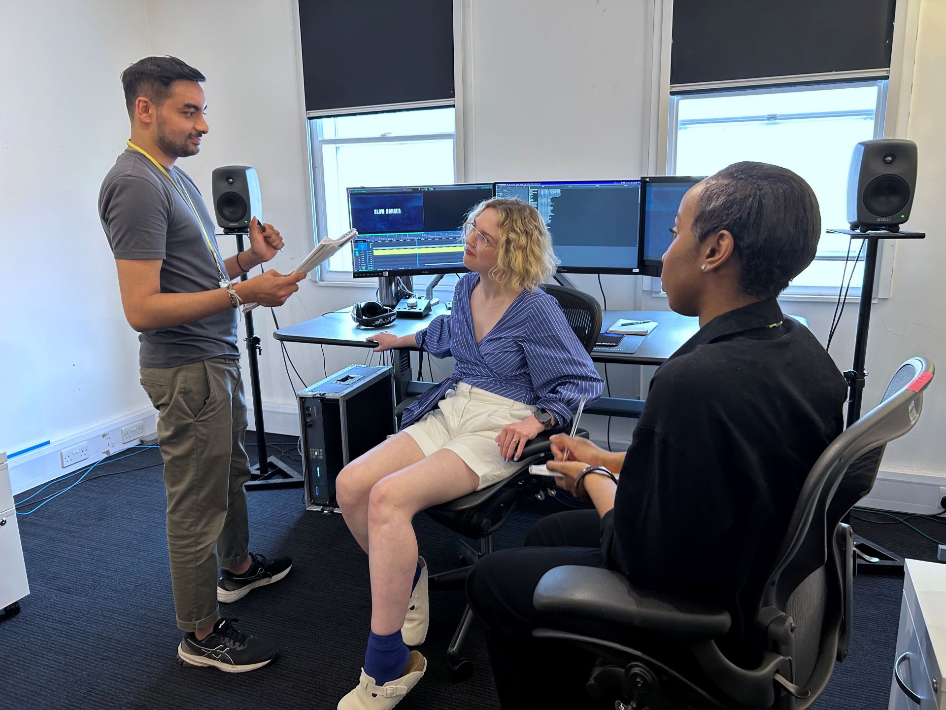 A person, stood, gives instructions to two other people, seated, in a room set up behind them with a set of computer monitors upon the desk and speakers on either size.