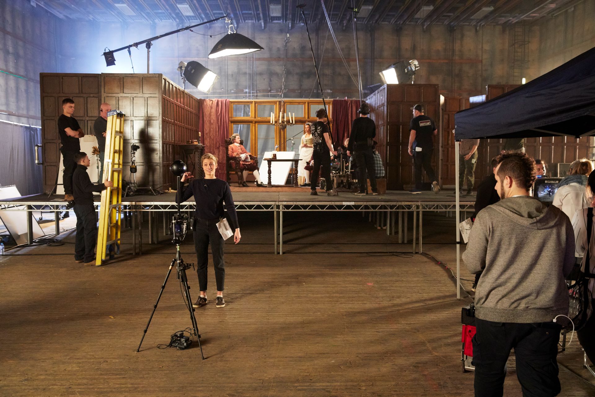 Behind the scenes image of crew working on a set with camera and lights being set up while actors in costume sit on a stage set up as a living room