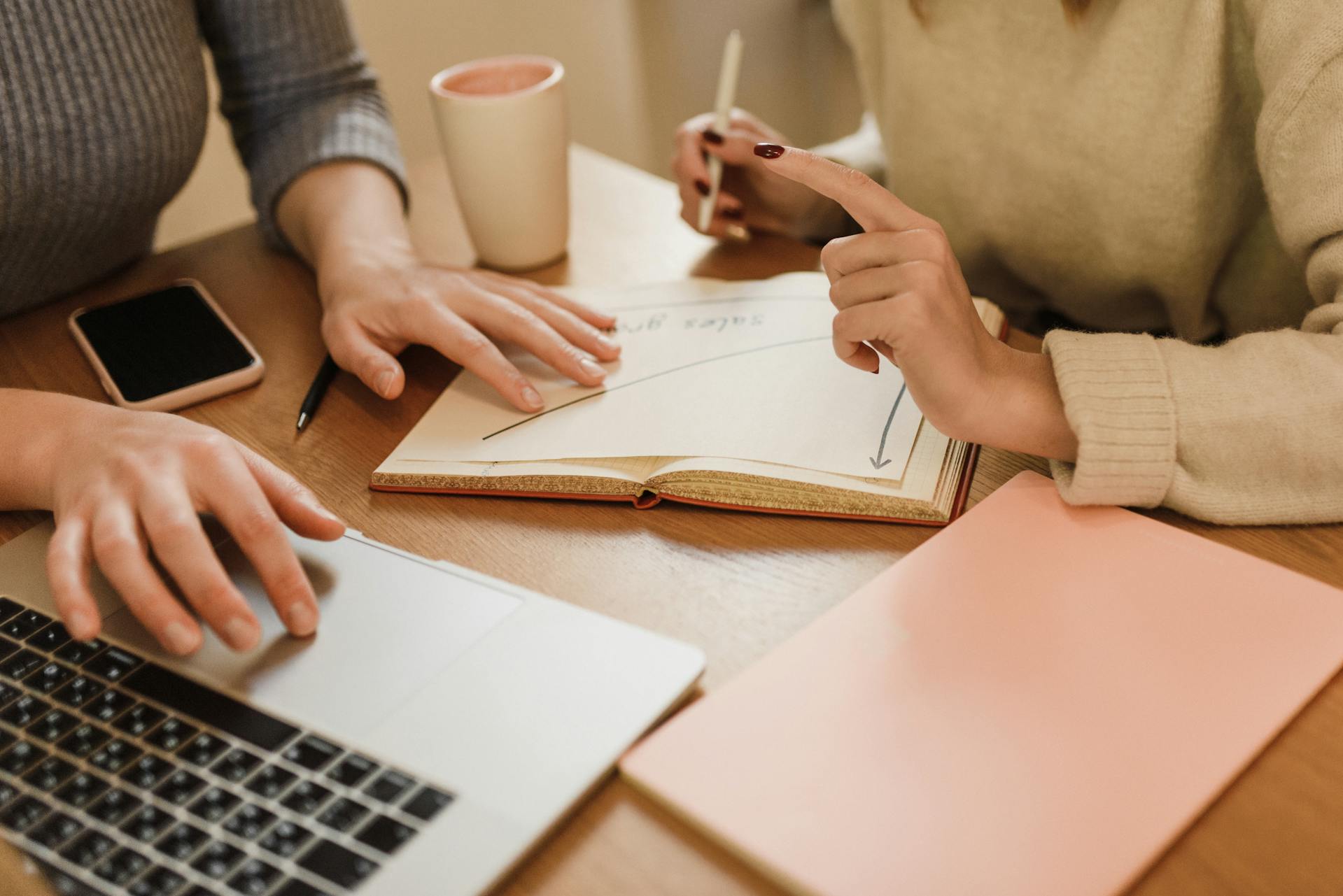 Two people sit at a desk with laptops and notebooks between them