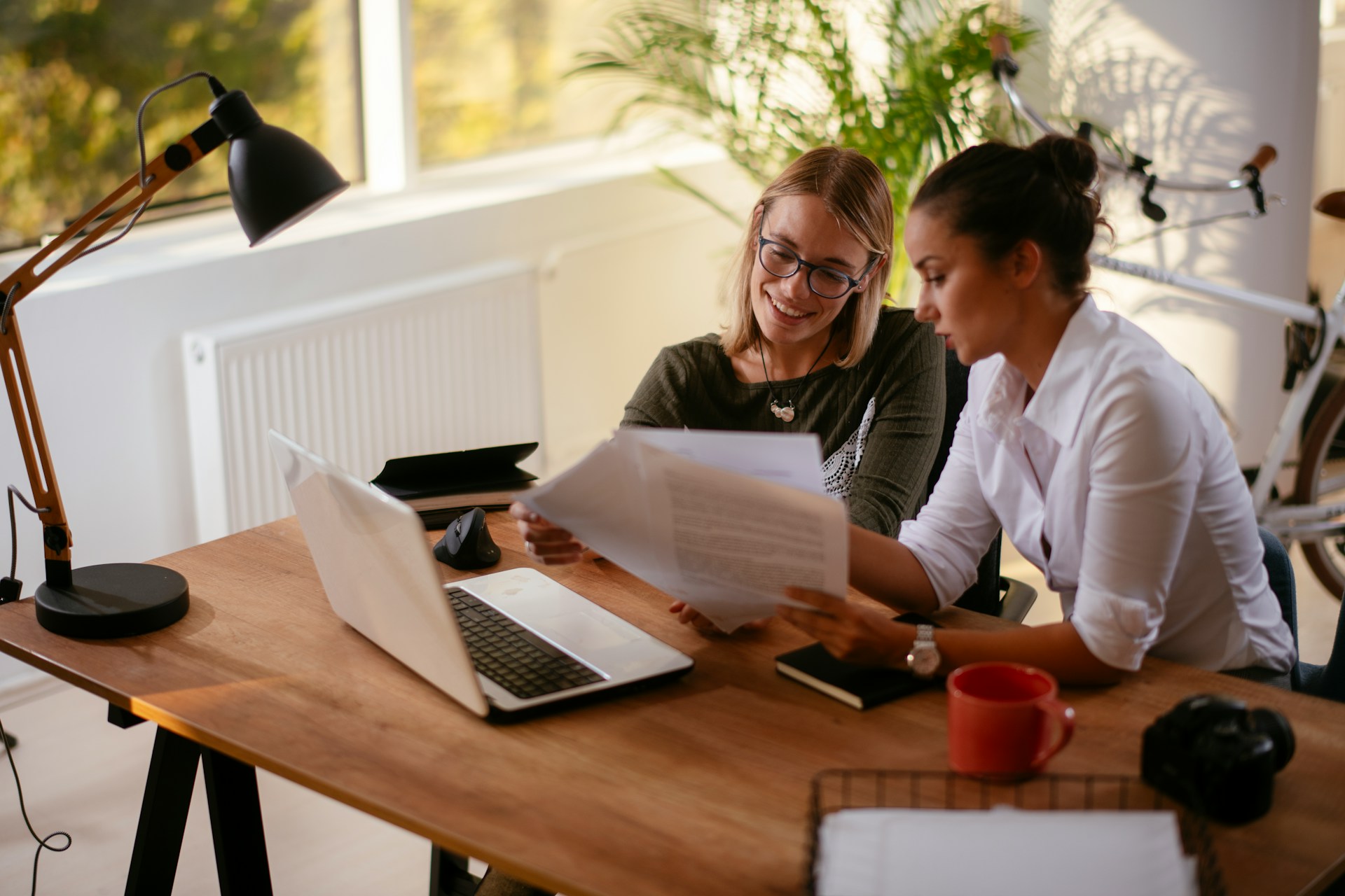 Two women sit at a desk with a laptop, looking through some papers