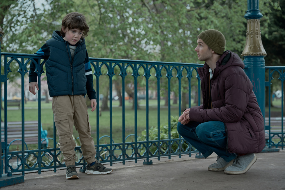 A man kneels down to speak to a child outside a park